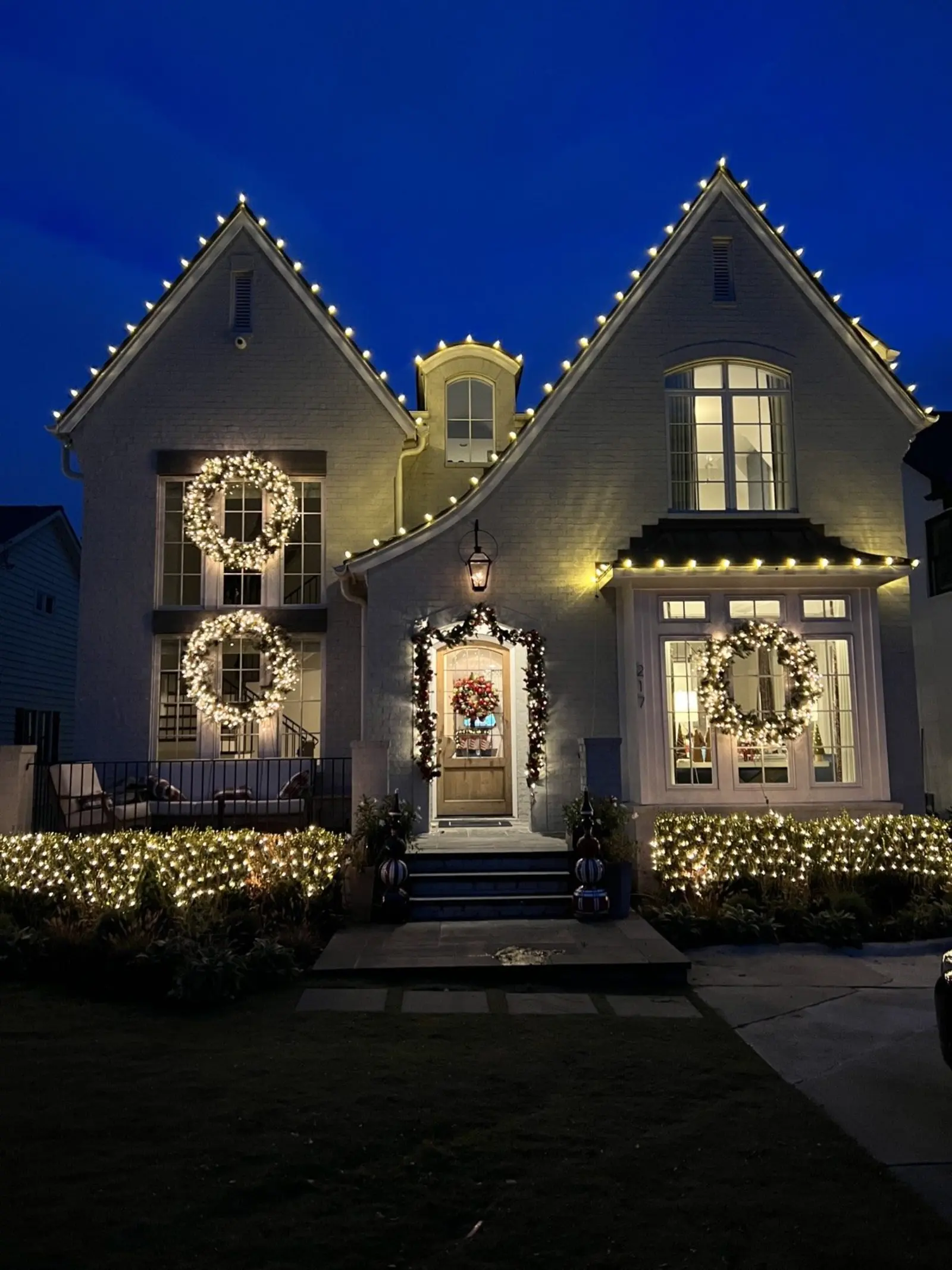 White brick home at dusk with warm white Christmas roofline lights, lighted wreaths and garland